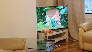 Uma sala de estar com televisão de tela plana sobre uma mesa de centro de vidro, sofás bege e uma janela com cortinas.