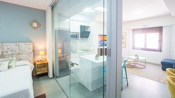 A modern hotel room with a glass partition separating the bedroom from the kitchenette.