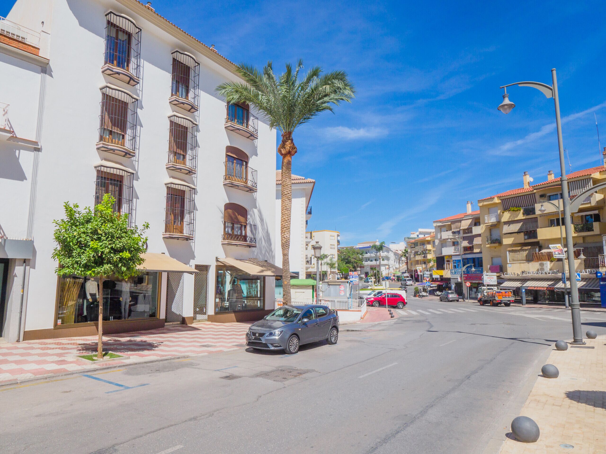 A street view with a building featuring multiple balconies, a palm tree, and a parked car.
