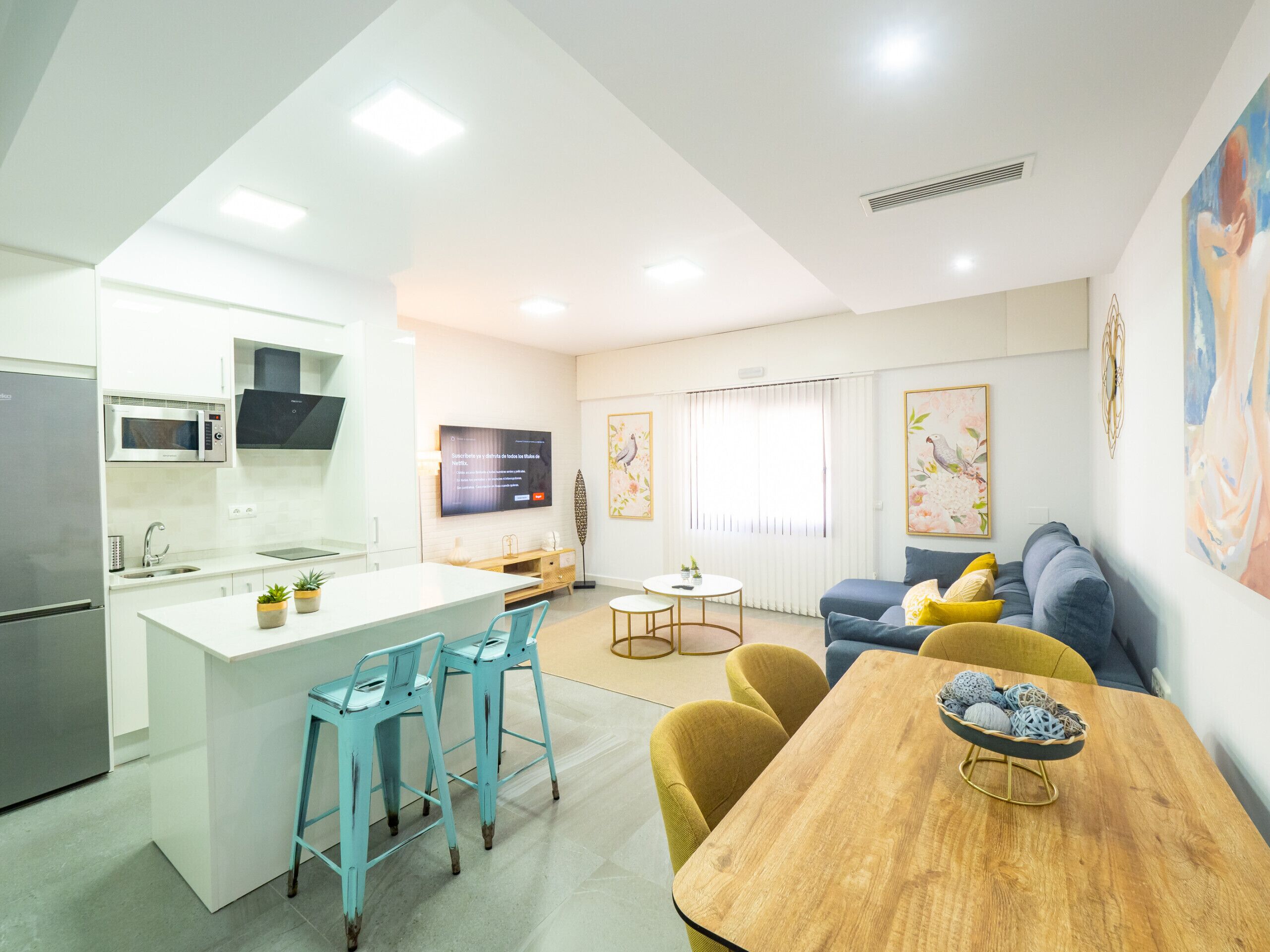 A modern kitchen with a central island, bar stools, a dining table with a bowl of decorative items, a sofa, and a TV mounted on the wall.