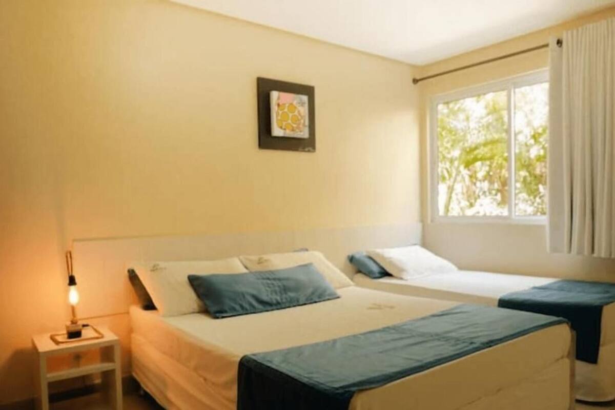 A hotel room with two beds, a bedside table, a lamp, a window with curtains, and a framed picture on the wall.