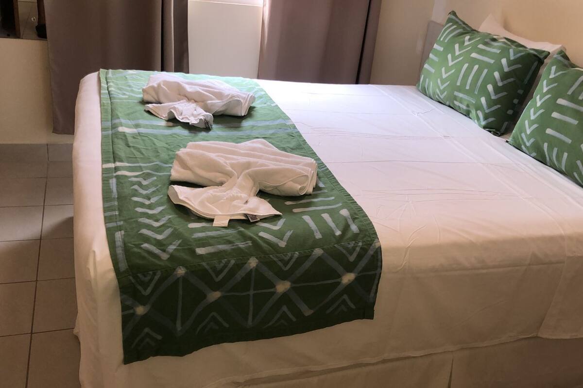 A neatly made bed with a green patterned bedspread and matching pillows, a white blanket, and a folded white towel on the bed.