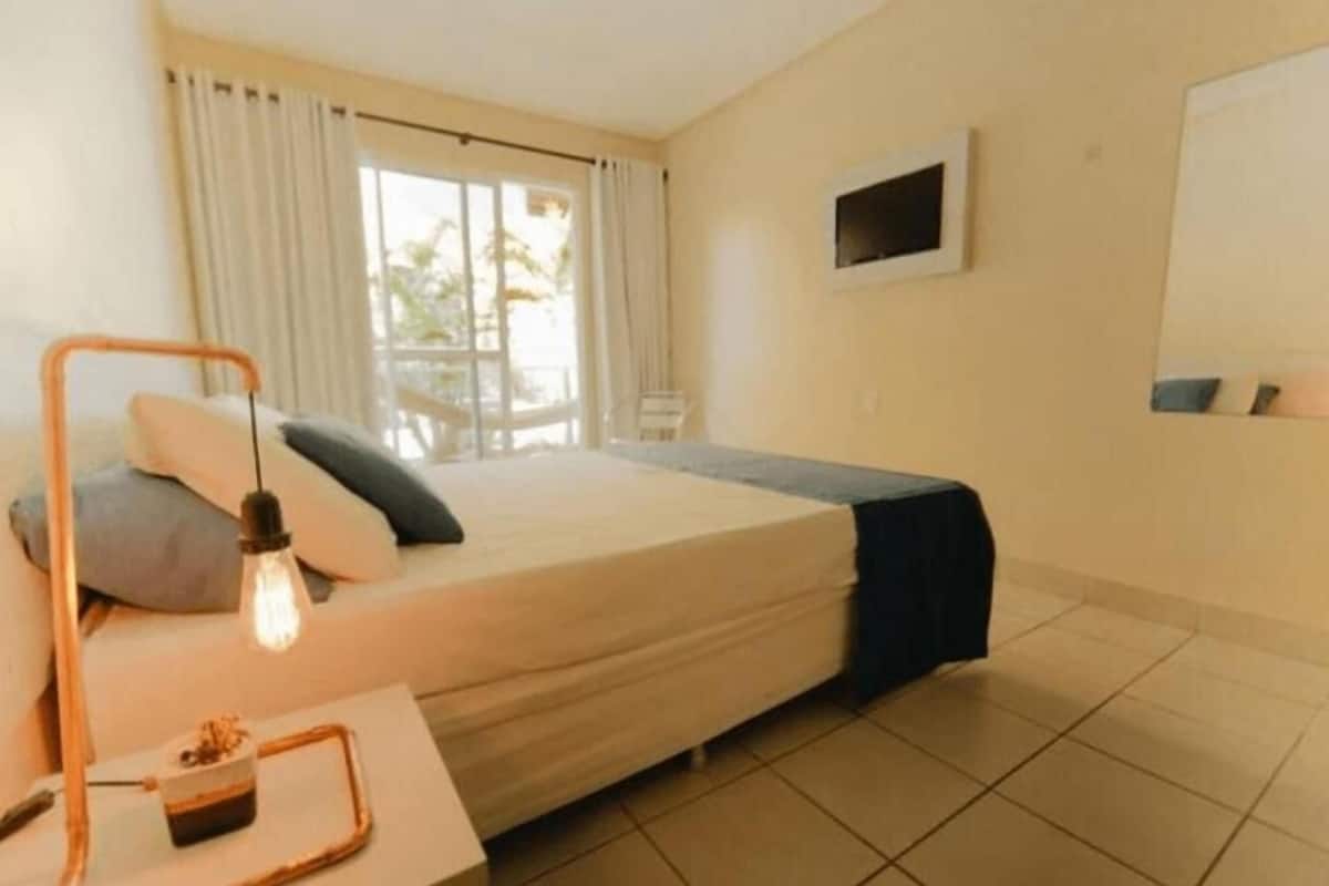 A hotel room with a bed, a bedside table with a lamp, a small table with a tray, a TV mounted on the wall, and a window with curtains.