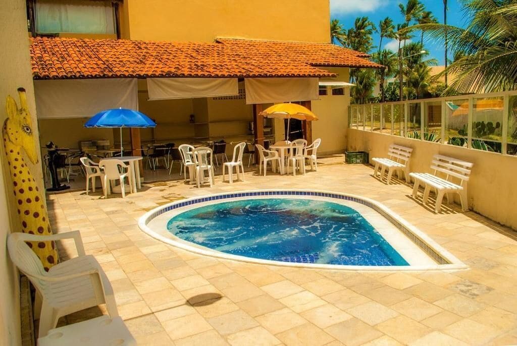 Una piscina con suelo de baldosas, rodeada de sillas y mesas de plástico blanco, y sombrillas que dan sombra.