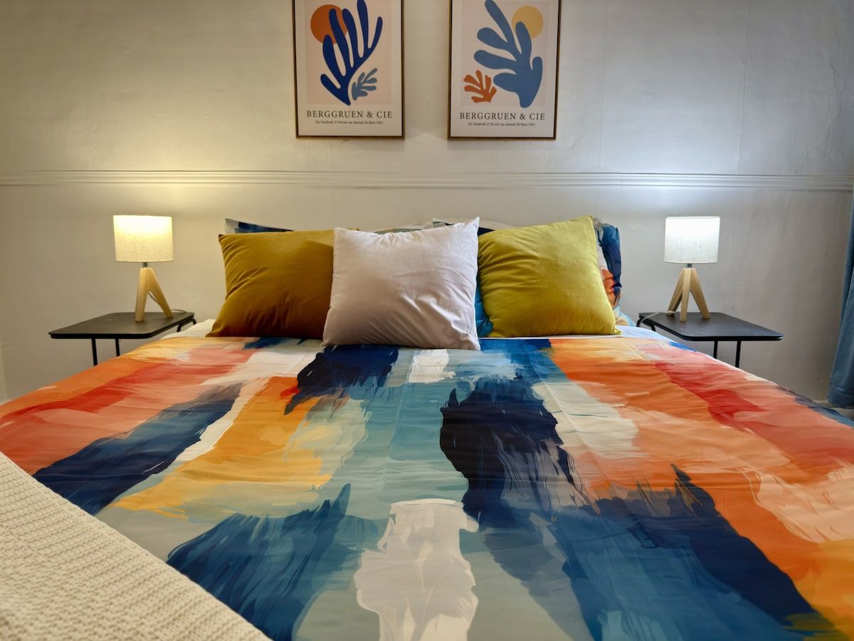 A bed with a colorful abstract bedspread, two bedside tables with lamps, and two framed artworks on the wall.