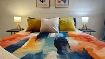A bed with a colorful abstract bedspread, two bedside tables with lamps, and two framed artworks on the wall.