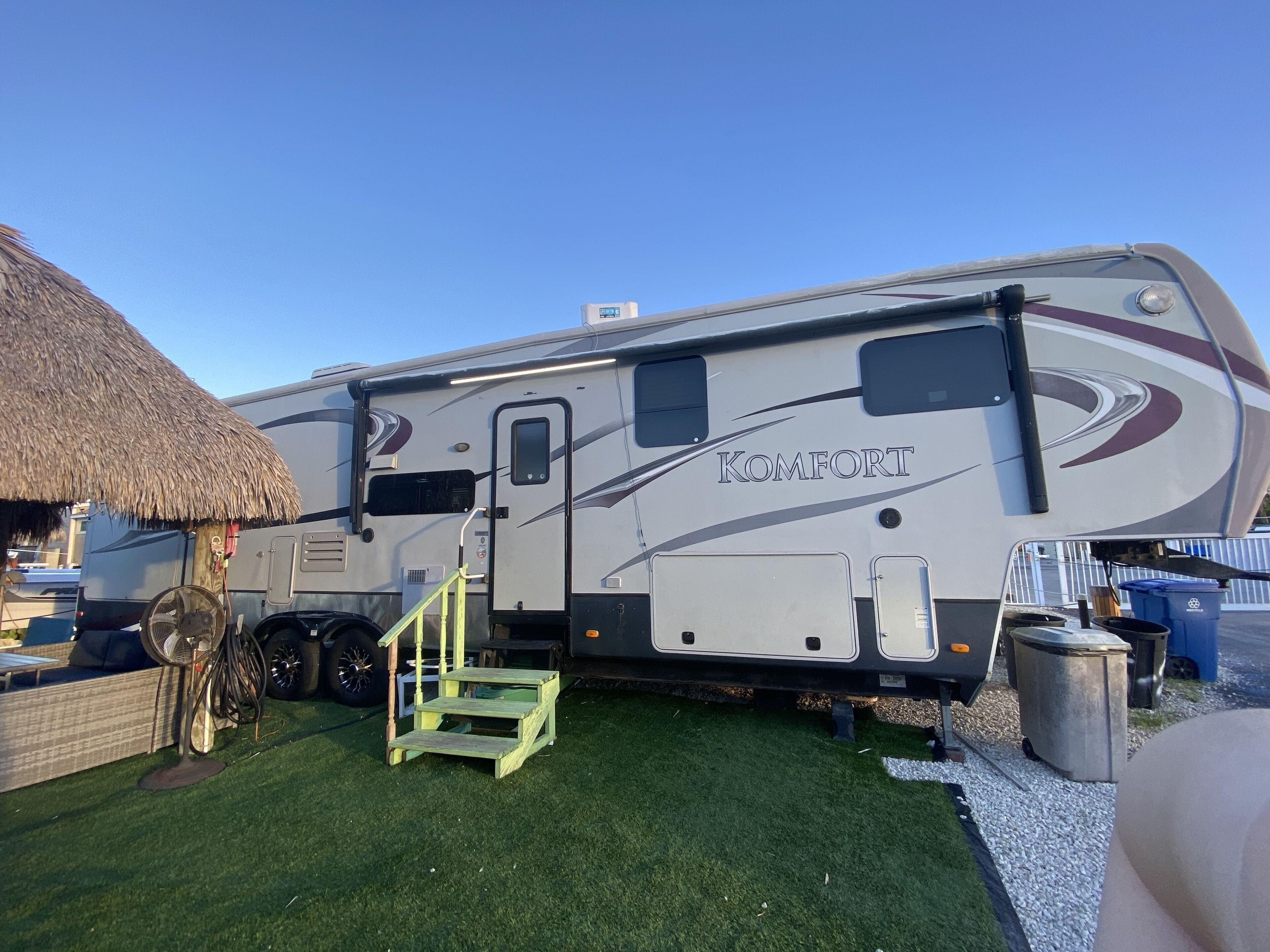 A large RV with the brand name 'Komfort' on the side, a small wooden step, and a thatched structure nearby.