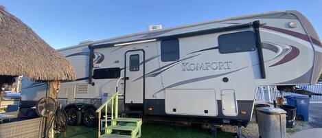 A large RV with the brand name 'Komfort' on the side, a small wooden step, and a thatched structure nearby.