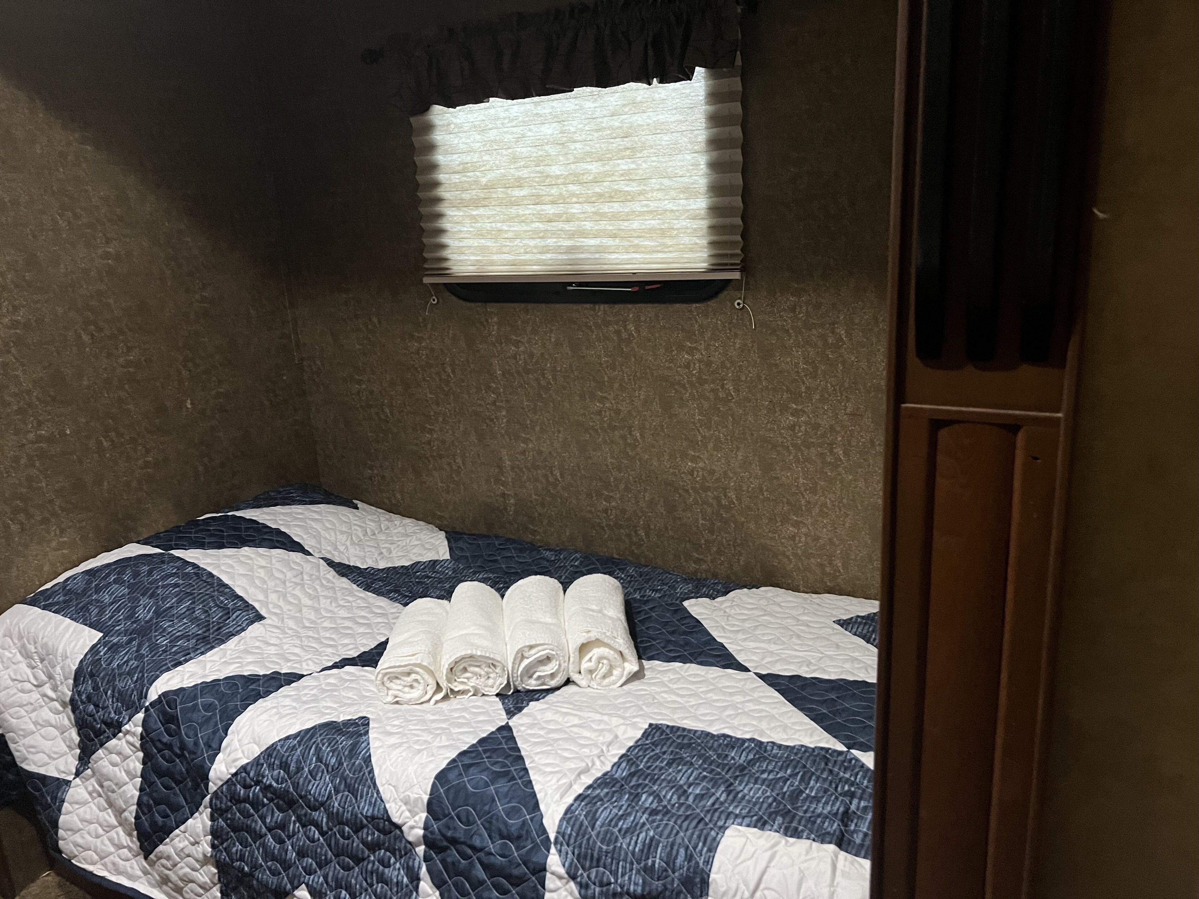 A small, single-bed room with a quilted bedspread, a window with blinds, and a wooden door.