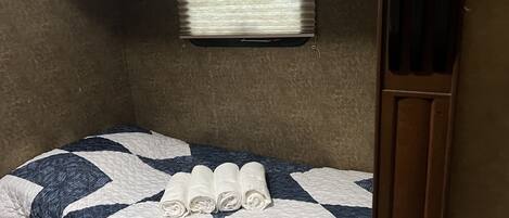 A small, single-bed room with a quilted bedspread, a window with blinds, and a wooden door.