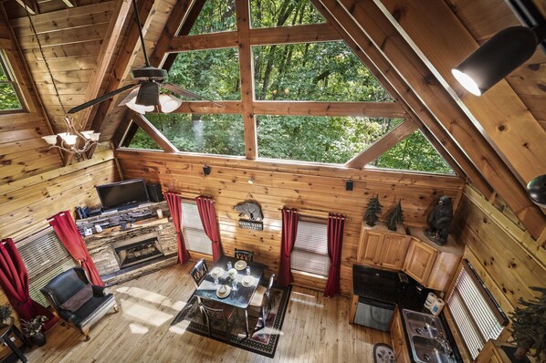Een chalet met een eethoek, een open haard, een televisie en grote ramen met uitzicht op een bos.