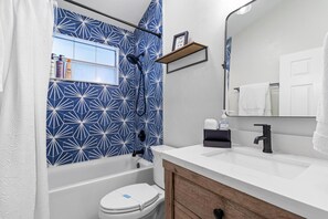 Un baño moderno con un salpicadero de azulejos azules geométricos, una bañera blanca, un inodoro blanco y un lavabo blanco con grifo negro, además de un mueble de tocador de madera.