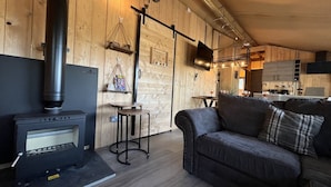 A cozy living room with a fireplace, wooden walls, a sofa, and a kitchen area in the background. - Lion King Safari Tent (Tenby)