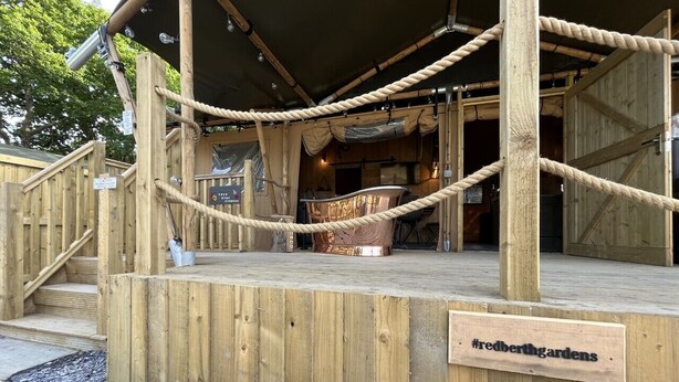 A wooden deck with a rope barrier, a sign reading "#redberthardens", and a view into a building with a covered area. - Lion King Safari Tent (Tenby)
