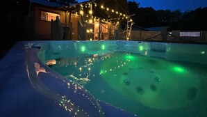 A hot tub with green lighting, a covered outdoor area with string lights, and a building in the background. - Lion King Safari Tent (Tenby)