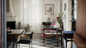 Ein gemütliches Wohnzimmer mit Kamin, einem Esstisch und einer Couch. Der Boden ist kariert und ein Fenster mit durchsichtigen Vorhängen.