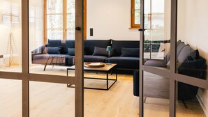 A modern living room with a dark-colored sofa, a coffee table, and a floor lamp. Large windows allow natural light to fill the space. - Light-flooded dream home between Chiemsee and the Alps (Traunstein)