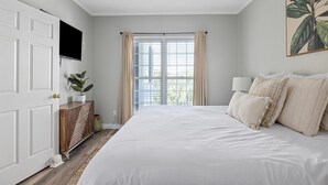 A bedroom with a large bed, a TV mounted on the wall, a window with curtains, and a ceiling fan. - Pet-friendly oceanfront condo steps from beach & center street (Folly Beach)