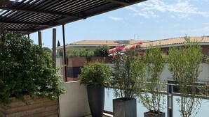 Un'area salotto all'aperto riparata, con tavolo e sedie, circondata da piante in vaso e vista su un edificio dall'altra parte dell'acqua.