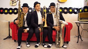 Vier Musiker sitzen auf einer roten Couch, mit Musikinstrumenten, einer Gitarre und einem Plattenspieler im Hintergrund.