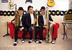 Cuatro músicos sentados en un sofá rojo, con instrumentos musicales, una guitarra y un tocadiscos al fondo.