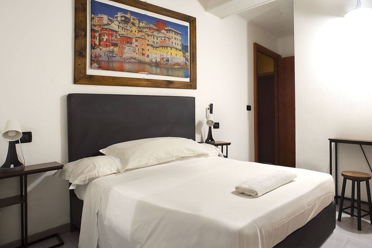 A hotel room with a bed, a nightstand, a lamp, a painting on the wall, and a small table with stools.