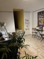 Huone, jossa on valkoinen tiiliseinä, pöytä, jossa on lamppu, ruukkukasvi ja seinä, johon on kirjoitettu teksti: ”Pink Floyd The Wall”.