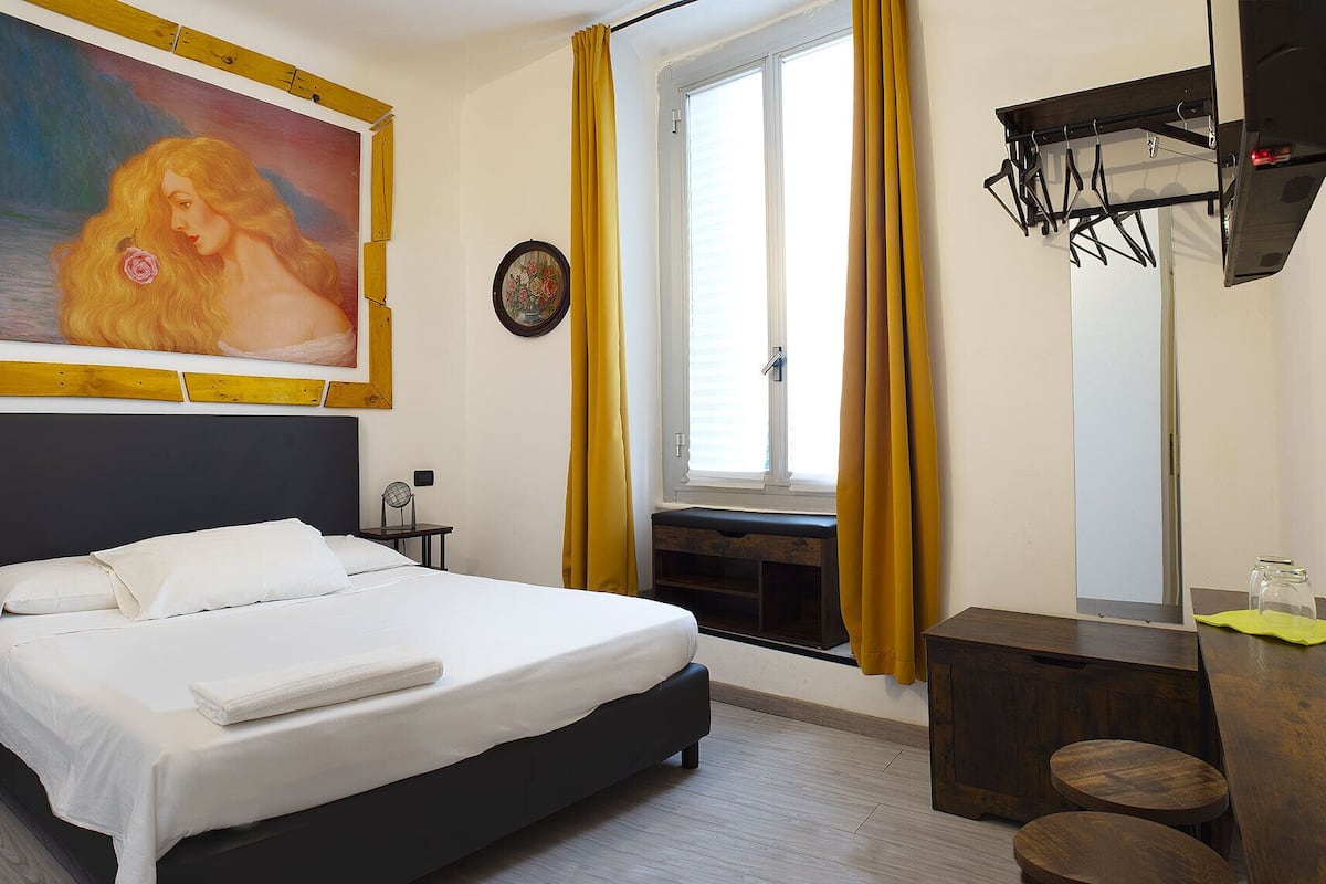A hotel room with a bed, a painting, a window with curtains, a bedside table, and a mirror.