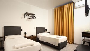 Ein Hotelzimmer mit zwei Betten, einem Holznachttisch, einem Schreibtisch und einem an der Wand befestigten Wäscheständer.