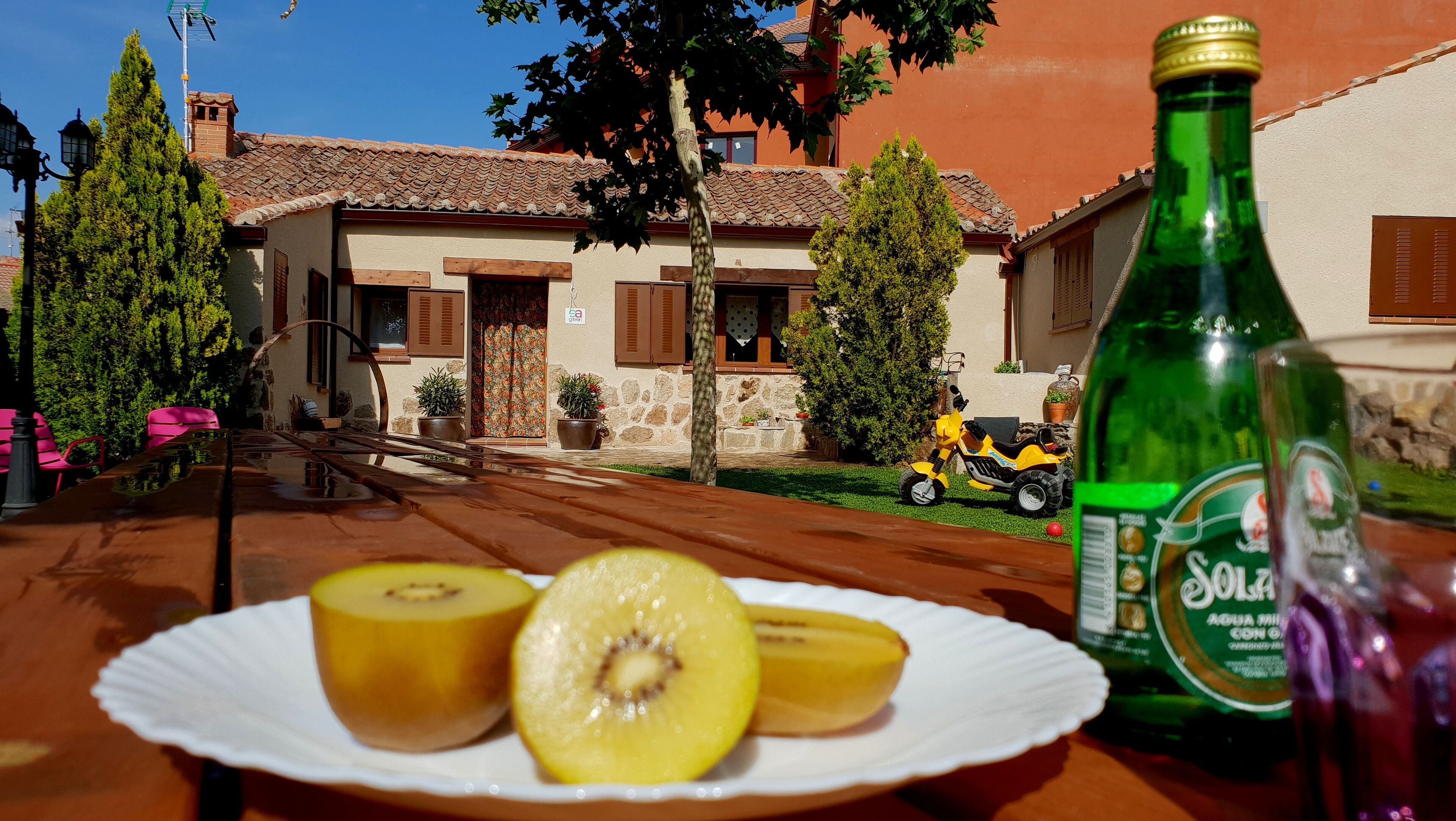 A table with a plate of fruit and a bottle of Solz beer in front of a house with a tiled roof and stone walls.