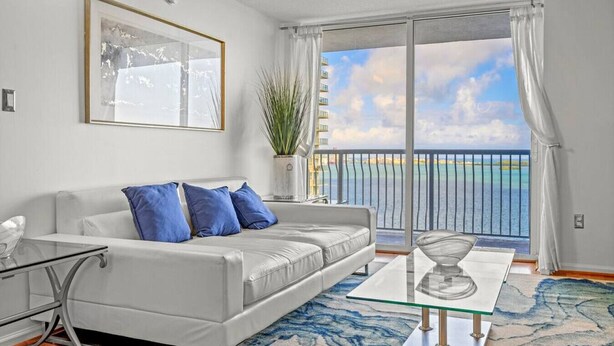 Una sala de estar moderna con un sofá blanco, cojines azules, una mesa de centro de vidrio y vistas al mar desde un balcón.