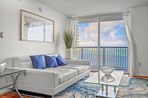 A modern living room with a white sofa, blue cushions, a glass coffee table, and a view of the sea through a balcony.