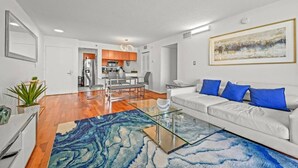 Una sala de estar moderna con una mesa de centro de vidrio, sofá blanco con cojines azules y una cocina al fondo.