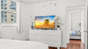 Un dormitorio con cama, cómoda con televisor mostrando una escena de playa, ventana con cortinas y vista de edificios exteriores.