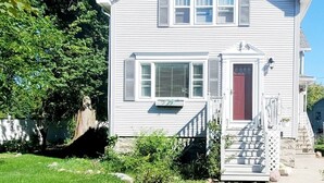 A two-story house with a red door, white siding, and a small porch with steps. - Spacious|Full Kitchen|Firepit|Office (Alpena)