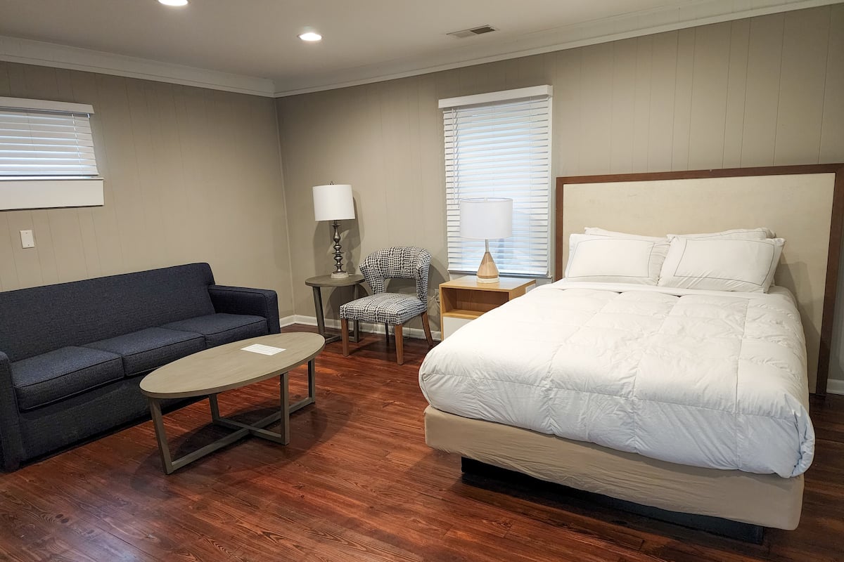 A hotel room with a large bed, a sofa, a chair, a small table, and two lamps.