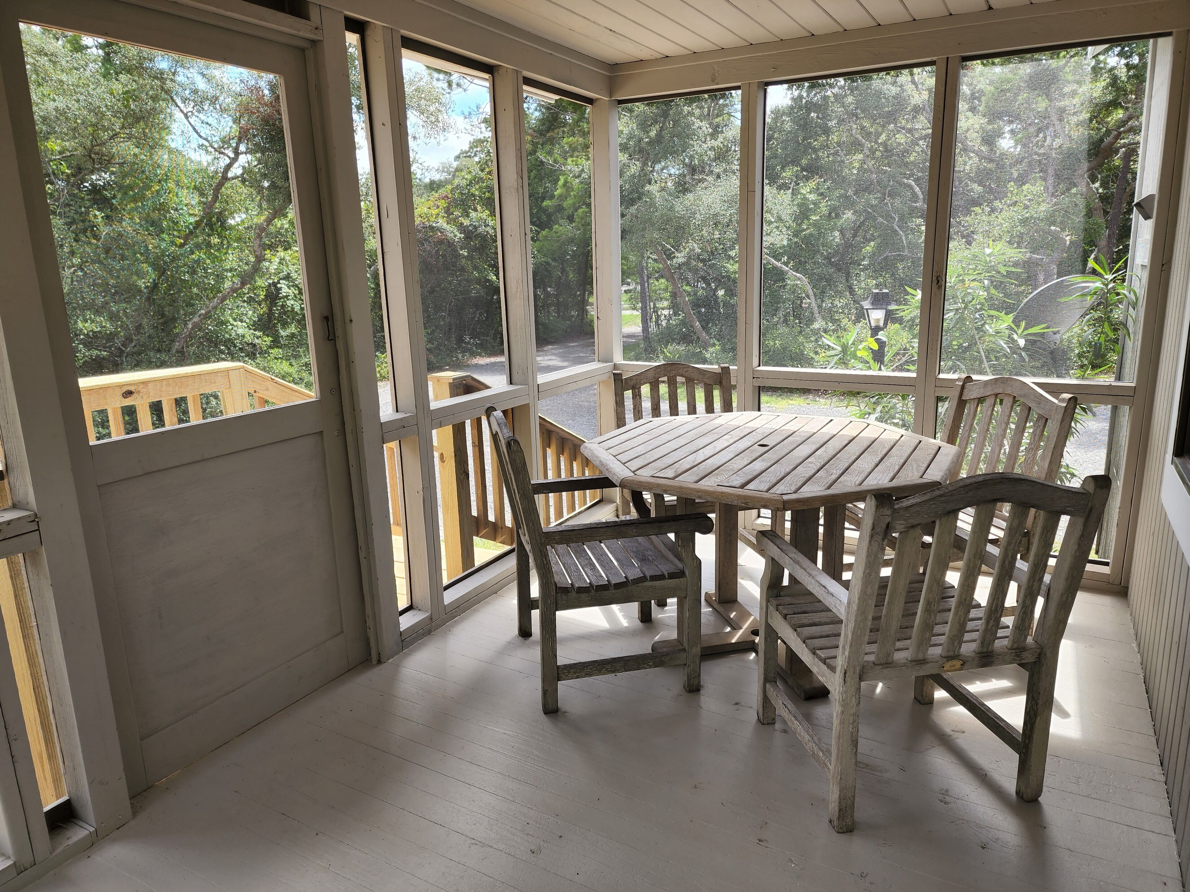 Een overdekt terras met een houten tafel en stoelen, grote ramen en uitzicht op de omliggende groene omgeving.