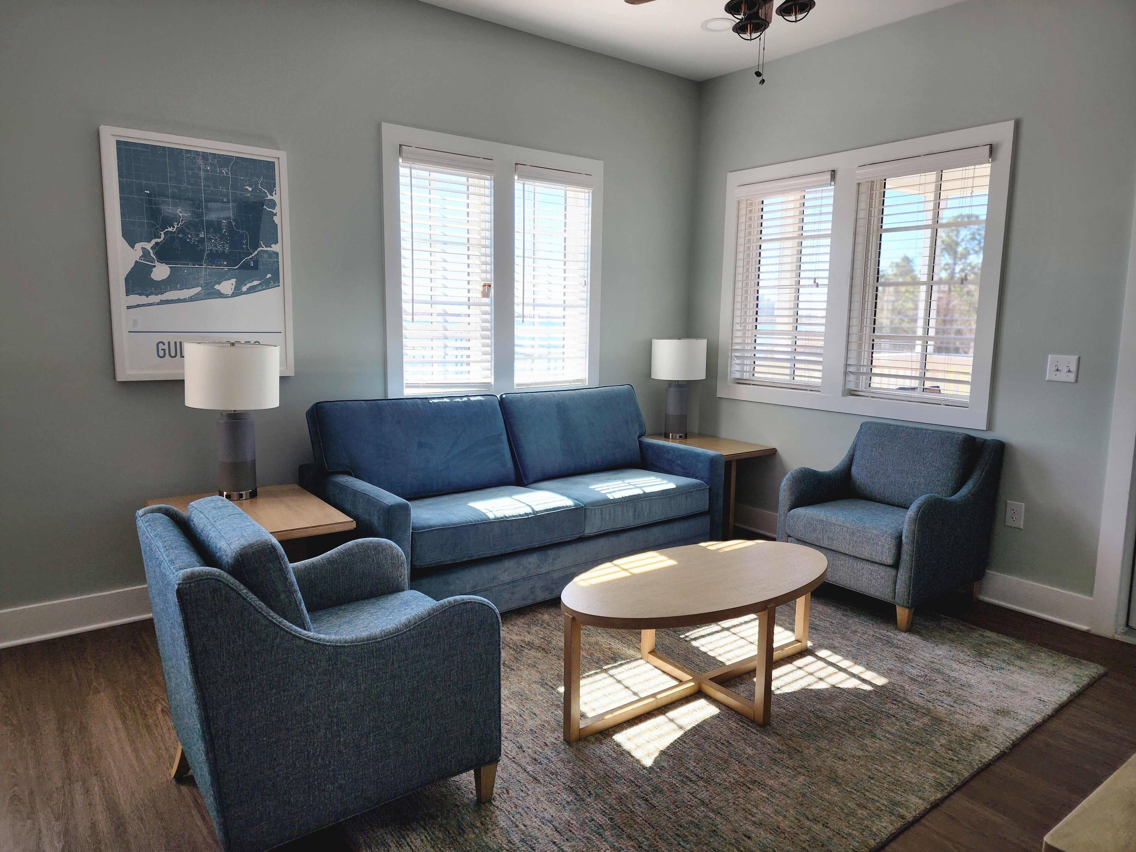 A living room with a blue sofa, two armchairs, a coffee table, and a rug. There are two windows with blinds, a lamp on a side table, and a map on the wall.
