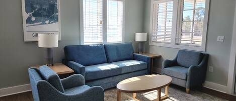 A living room with a blue sofa, two armchairs, a coffee table, and a rug. There are two windows with blinds, a lamp on a side table, and a map on the wall.