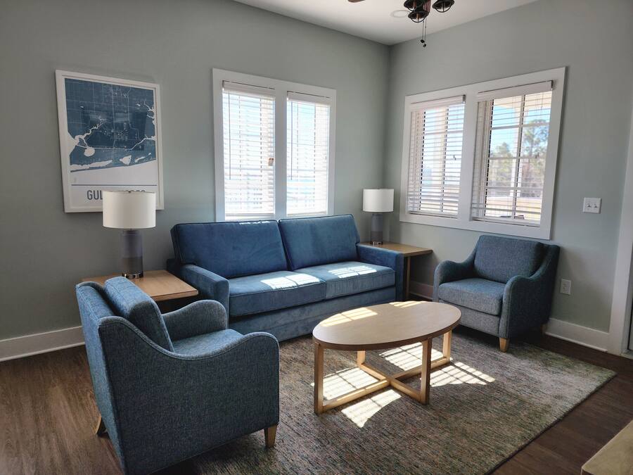 A living room with a blue sofa, two armchairs, a coffee table, and a rug. There are two windows with blinds, a lamp on a side table, and a map on the wall.