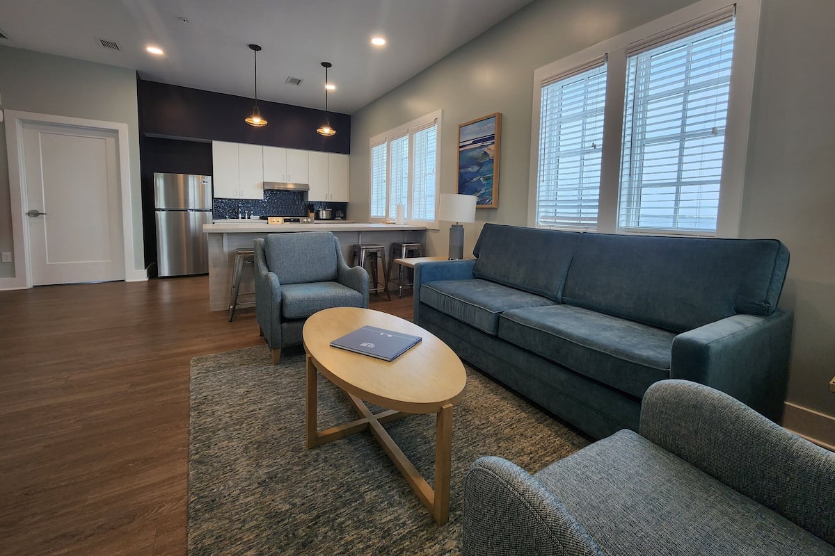 A modern living room with a blue sofa, a wooden coffee table, and a kitchen area with a stainless steel refrigerator and dark blue backsplash.