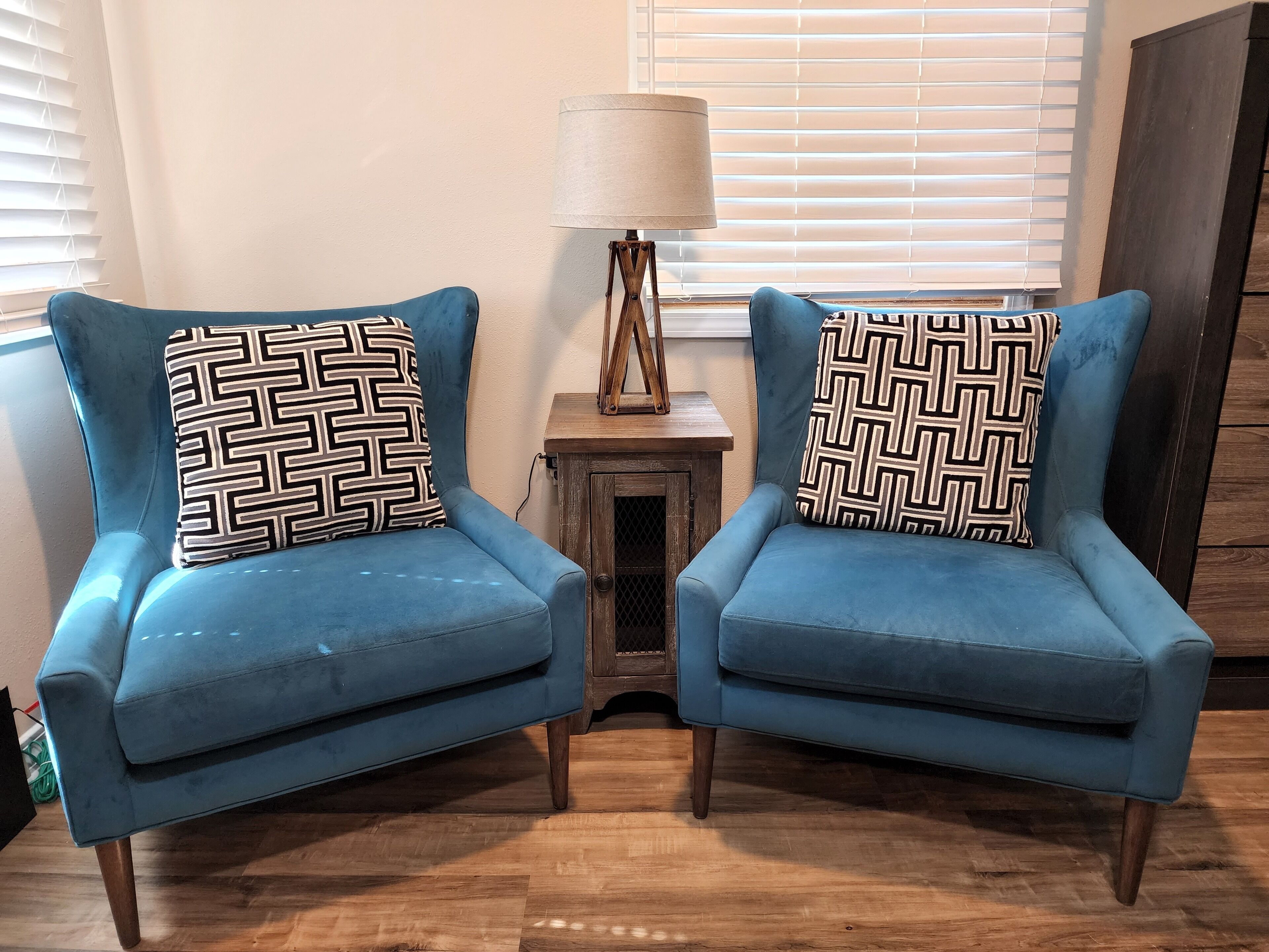Two blue armchairs with patterned cushions, a small wooden side table with a lamp, and a window with blinds.