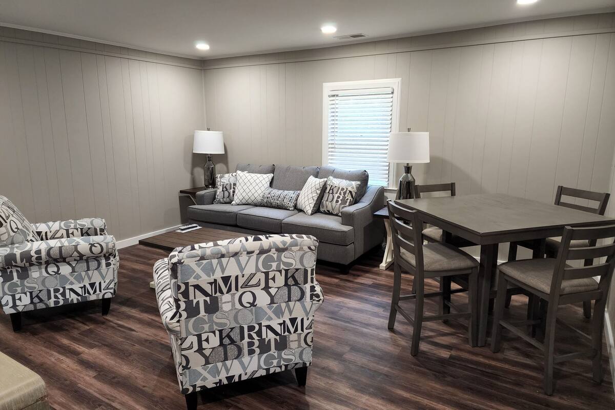 A living room with a grey sectional sofa, two armchairs with a letter pattern, a wooden dining table with chairs, and a window with blinds.