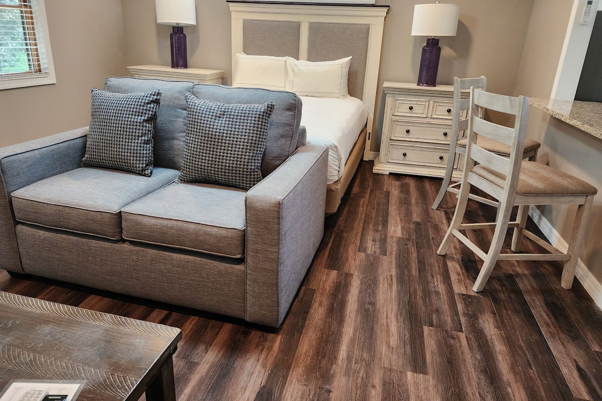 A compact bedroom with a grey sofa, a bed with white bedding, a wooden dresser, and a chair.