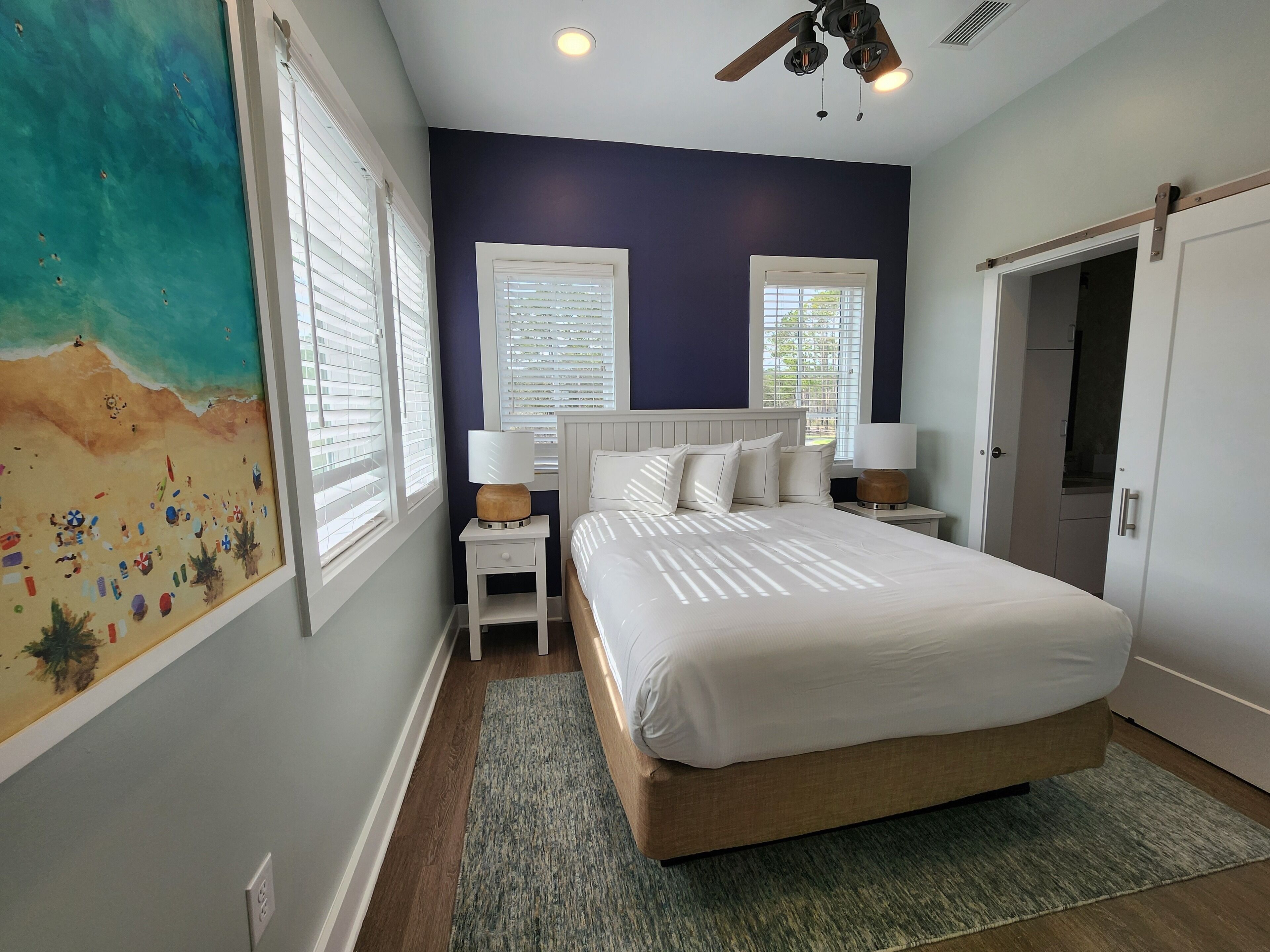 A bedroom with a bed, two bedside tables with lamps, a ceiling fan, and a beach-themed painting on the wall.