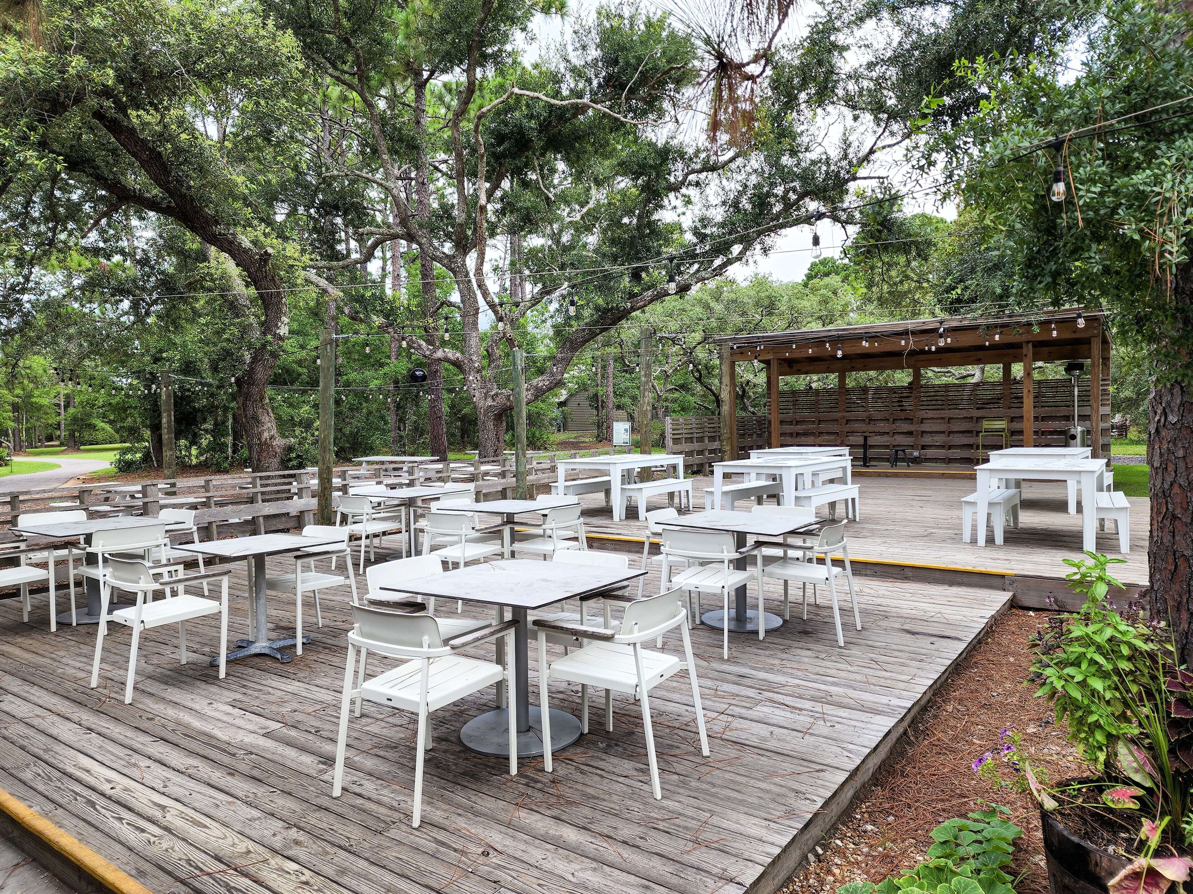 Un área de comedor al aire libre con mesas y sillas blancas, rodeada de árboles.
