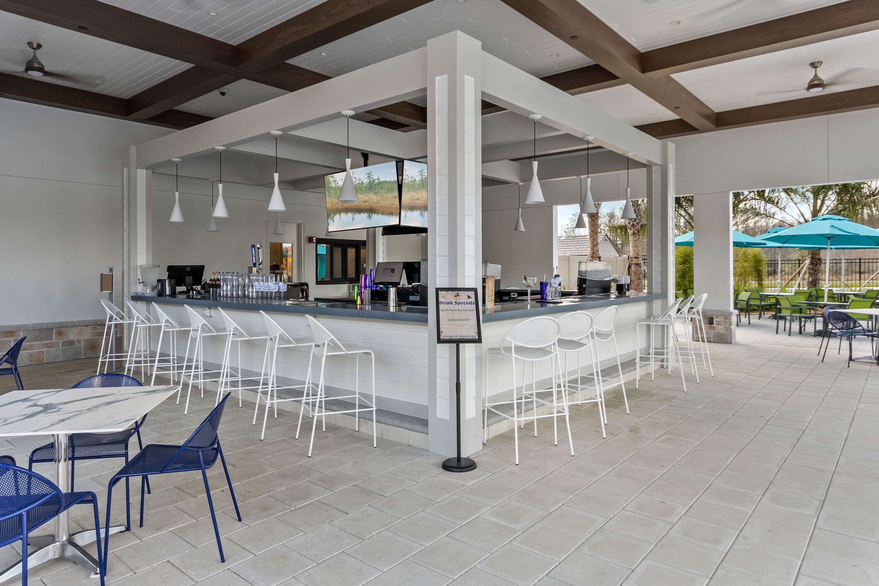 A spacious outdoor bar area with white bar stools and tables, a counter with a drink specialist sign, and hanging pendant lights. - NEW 9Bdrm Themed w Arcade near Disney & Universal (Kissimmee)