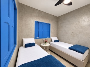 A room with two beds, a small round table, a ceiling fan, and a window with blue shutters. - Casa Marataizes (Marataizes)