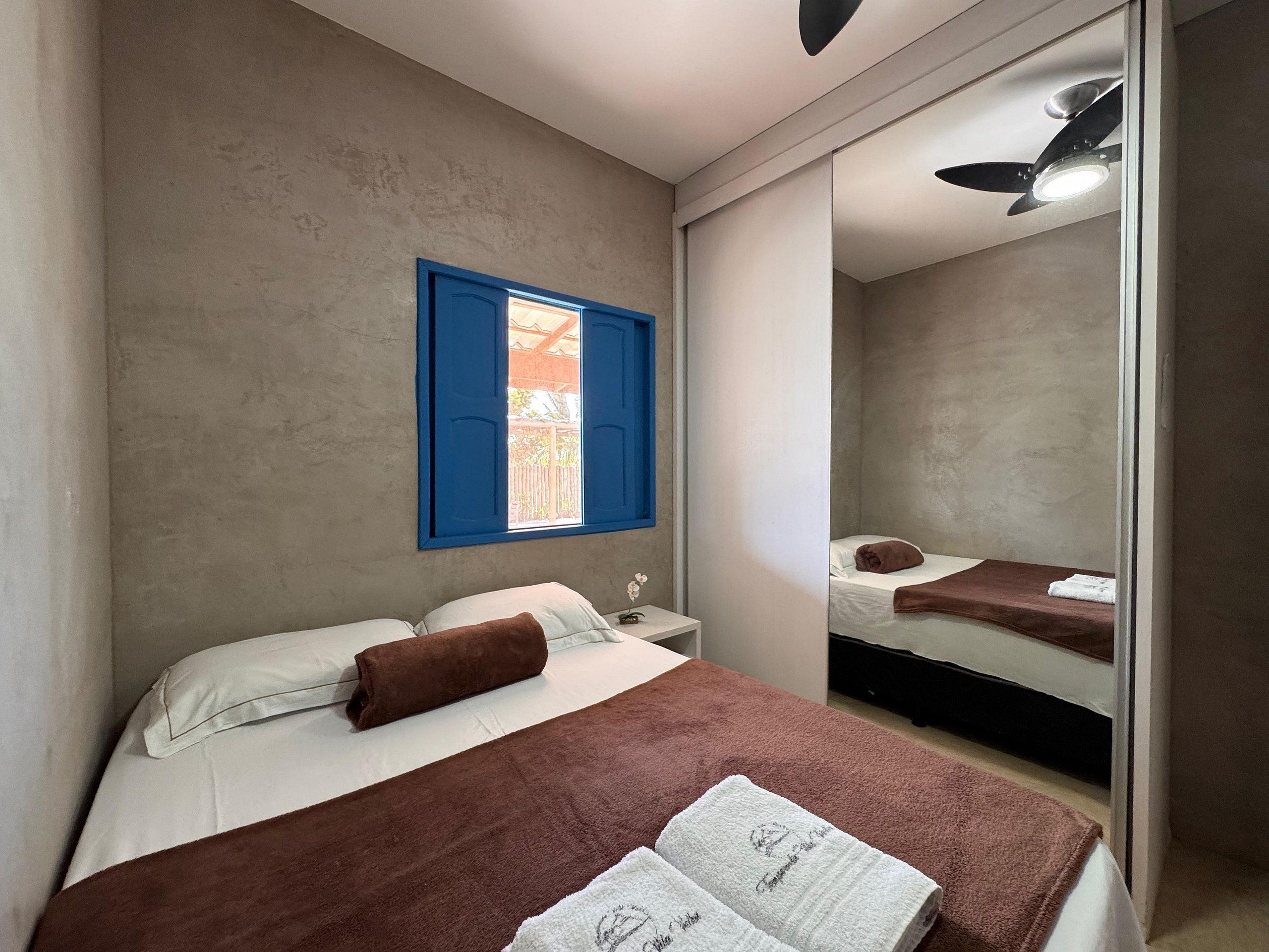 Quarto com cama, mesa de cabeceira, espelho e janela com persianas azuis.