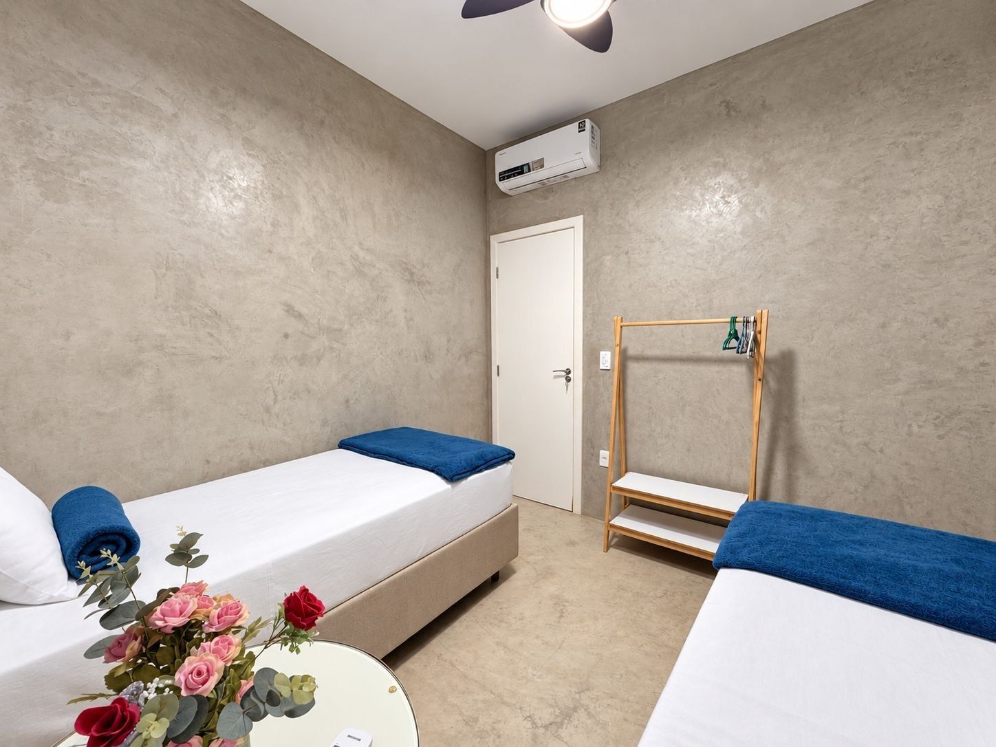 Quarto de hotel com duas camas, incluindo uma cama beliche de madeira, uma mesa redonda com um vaso de flores e um aparelho de ar condicionado na parede.